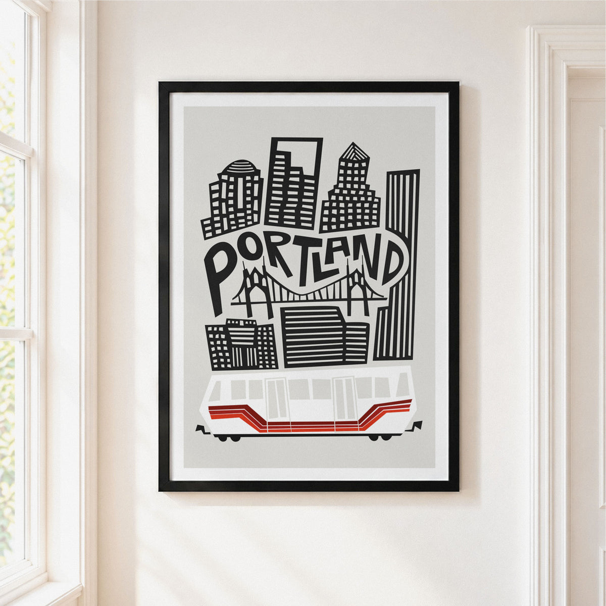 A framed Portland cityscape art print hangs on a white wall. The print features black and gray buildings and the word 'PORTLAND' with a red-and-white transit vehicle at the bottom.