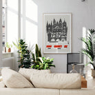 A framed art print featuring a black and white hand-drawn illustration of Prague's skyline with red accents, displayed on a light gray wall in a modern living room setting with a beige sofa and potted plants.