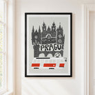 A framed art print featuring a stylized black and white illustration of Prague's skyline with the word 'PRAGUE' and a red-and-white tram, hanging on a light beige wall next to a window.