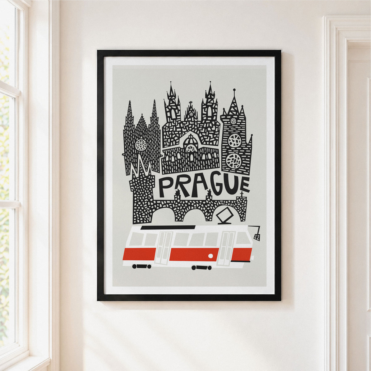 A framed art print featuring a stylized black and white illustration of Prague's skyline with the word 'PRAGUE' and a red-and-white tram, hanging on a light beige wall next to a window.
