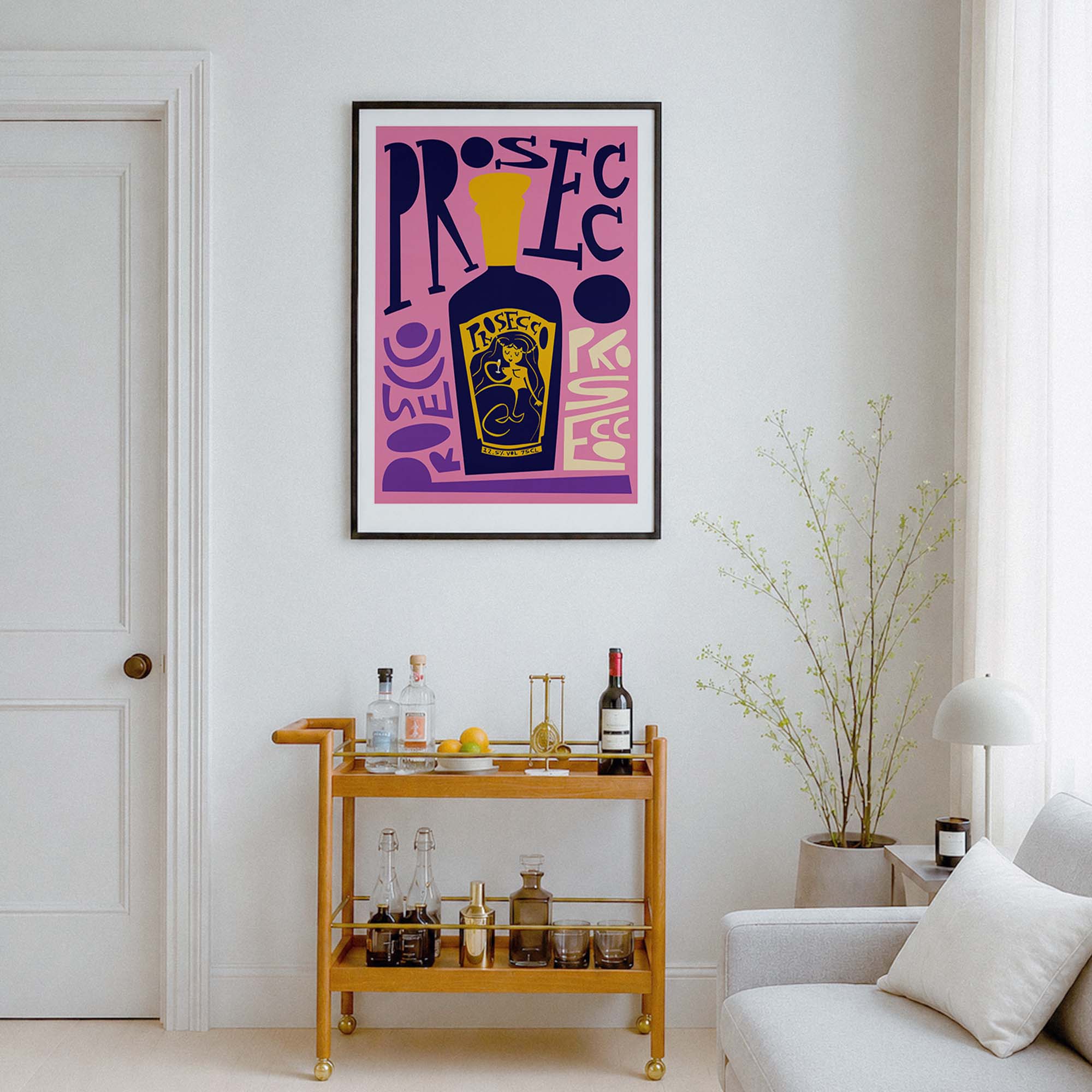 A framed pop-art print featuring a purple and pink background with a yellow bottle of prosecco illustration and stylized text, displayed on a white wall above a wooden bar cart in a modern living room setting.