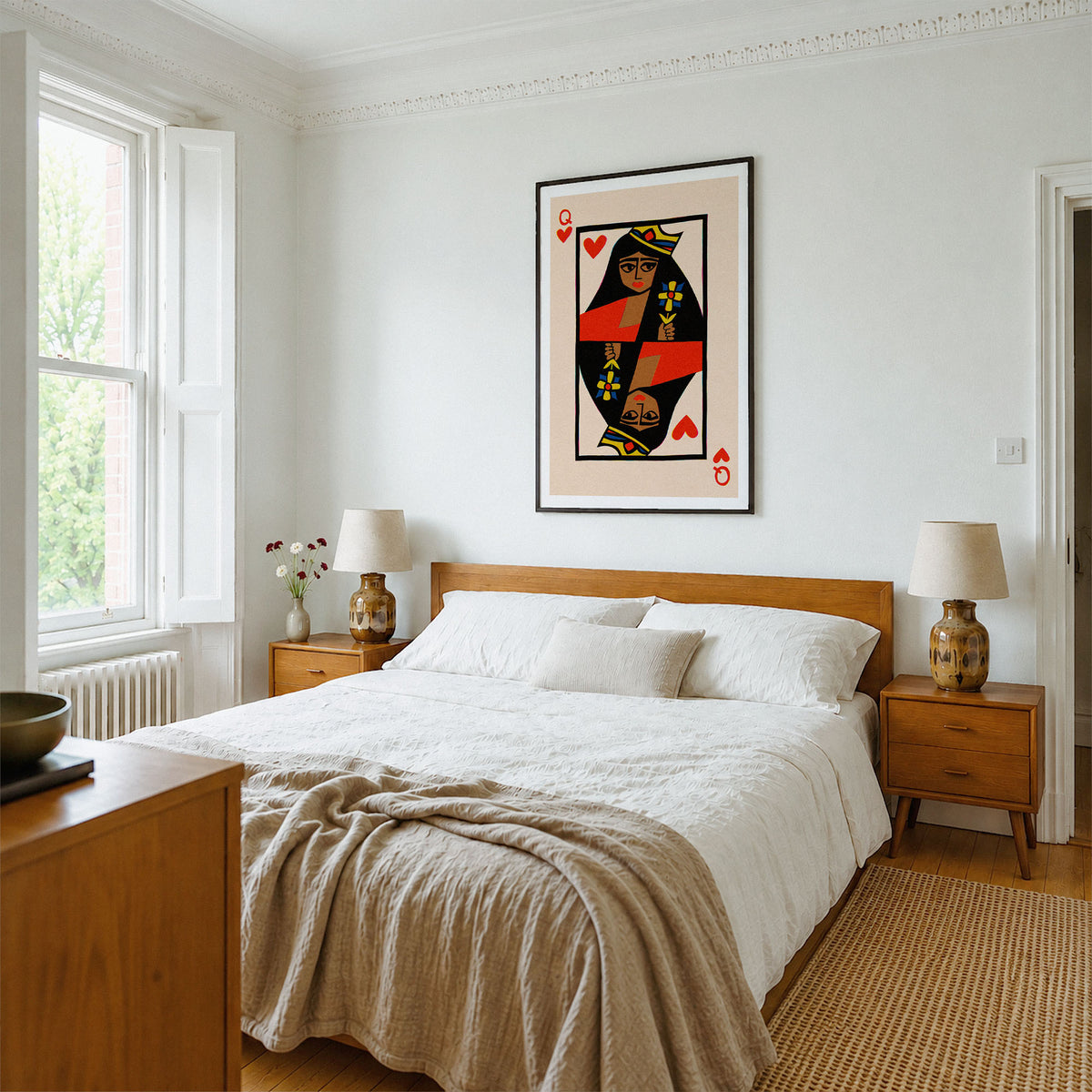 A styled bedroom featuring a large framed art print of a Queen of Hearts playing card hanging on the wall above a wooden bed. The room has white walls, a wooden headboard and nightstands, white bedding, and a beige textured throw blanket.