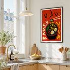 A framed vertical art print featuring a retro-style ramen illustration with the words 'RAMEN YUM' hangs on a white kitchen wall above a countertop with a sink, plants, and wooden cabinets.