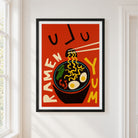 A framed retro-style ramen poster hanging on a white wall next to a window. The poster features a bold red background with a black bowl of ramen, yellow noodles, chopsticks, and the words 'RAMEN' and 'YUM' in cream and yellow lettering.