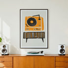 A framed art print featuring a retro orange and black record player illustration, displayed on a wall above a wooden credenza with speakers and a record turntable.