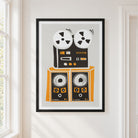 A framed art print featuring a retro graphic of a reel-to-reel tape machine and two speakers in black, orange, and white, displayed on a warm off-white wall next to a window.