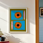 A framed abstract geometric art print hangs on a white wall. The print features two stacked blue rectangles with orange squares, black circles, olive green shapes, and pink vertical ovals.