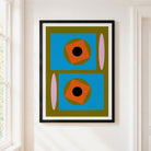 A framed abstract geometric art print hanging on a white wall next to a window. The print features a mid-century modern design with blue rectangles, orange rotated squares, olive green circles, pink vertical ovals, and black centers, all enclosed in a black frame with a white mat.