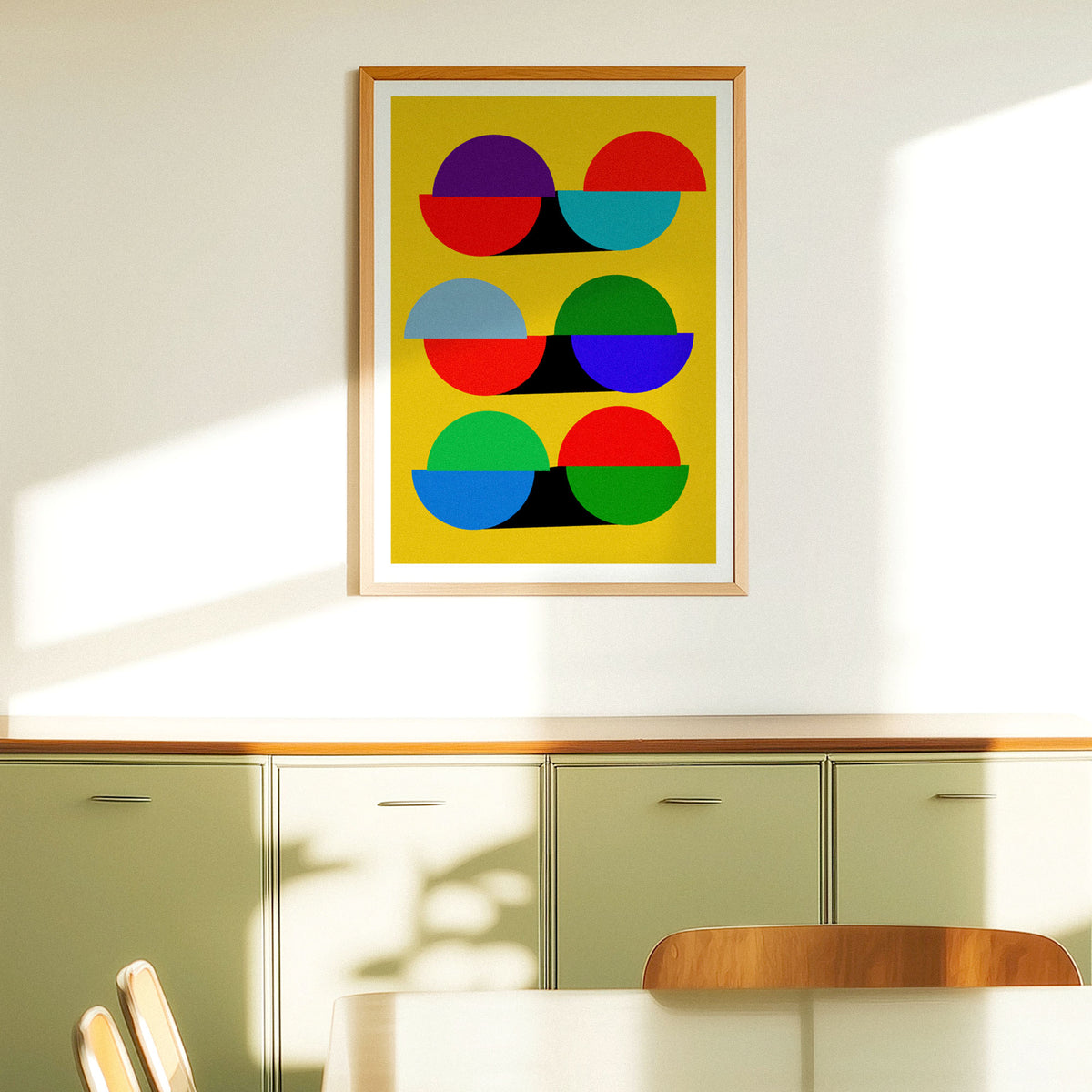 A framed abstract art print featuring colorful geometric circles on a yellow background, hanging on a white wall above a green cabinet in a modern room.