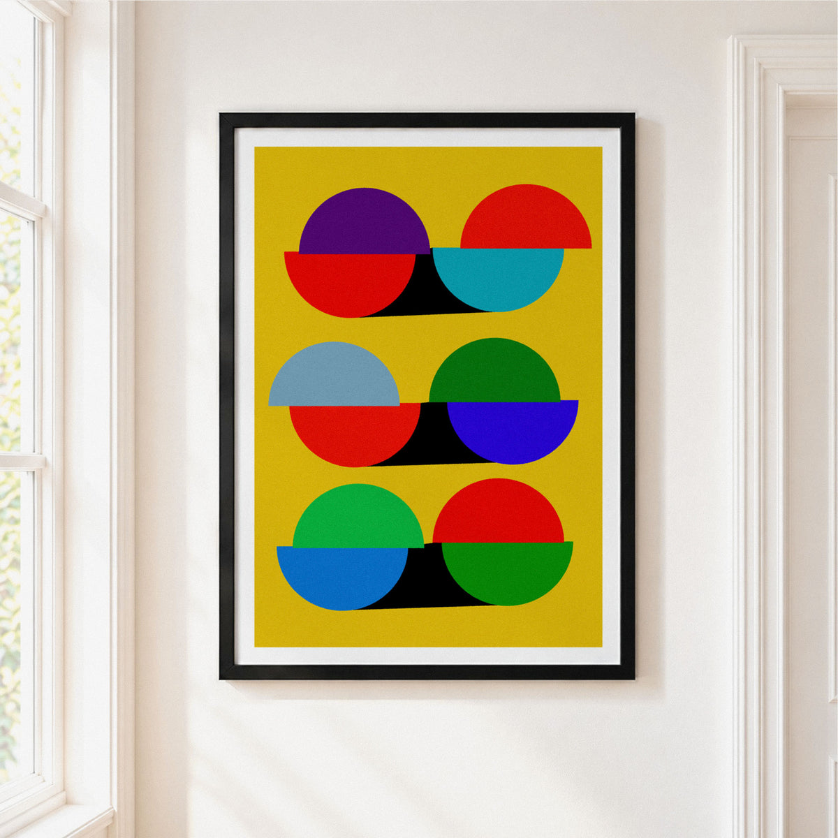 A framed abstract geometric art print featuring colorful semi-circles on a mustard yellow background, hung on a white wall next to a window.
