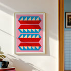 A framed art print featuring a mid-century modern geometric pattern of red and blue stylized shapes with cream triangles on a pink background, hung on a white wall next to a window.