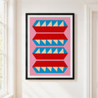 A framed art print featuring a mid-century modern geometric pattern of red and blue stylized shapes with cream triangles on a pink background, hung on a white wall next to a window.