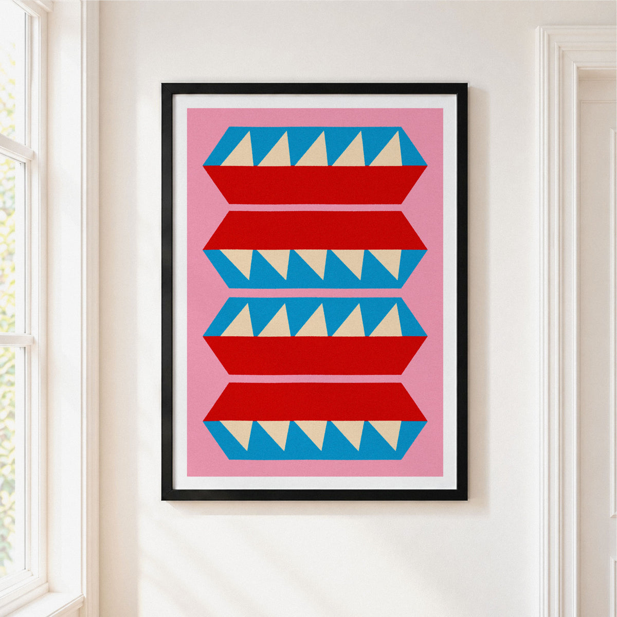 A framed art print featuring a mid-century modern geometric pattern of red and blue stylized shapes with cream triangles on a pink background, hung on a white wall next to a window.