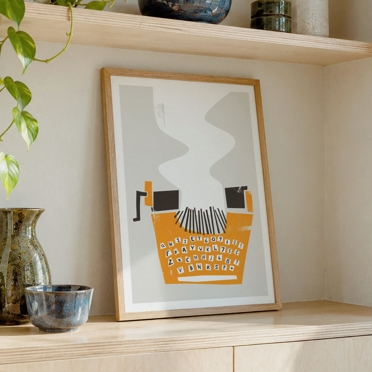 A framed abstract typewriter art print leaning on a light wood shelf, styled with ceramic vases and a trailing green plant in a warm, neutral interior setting.