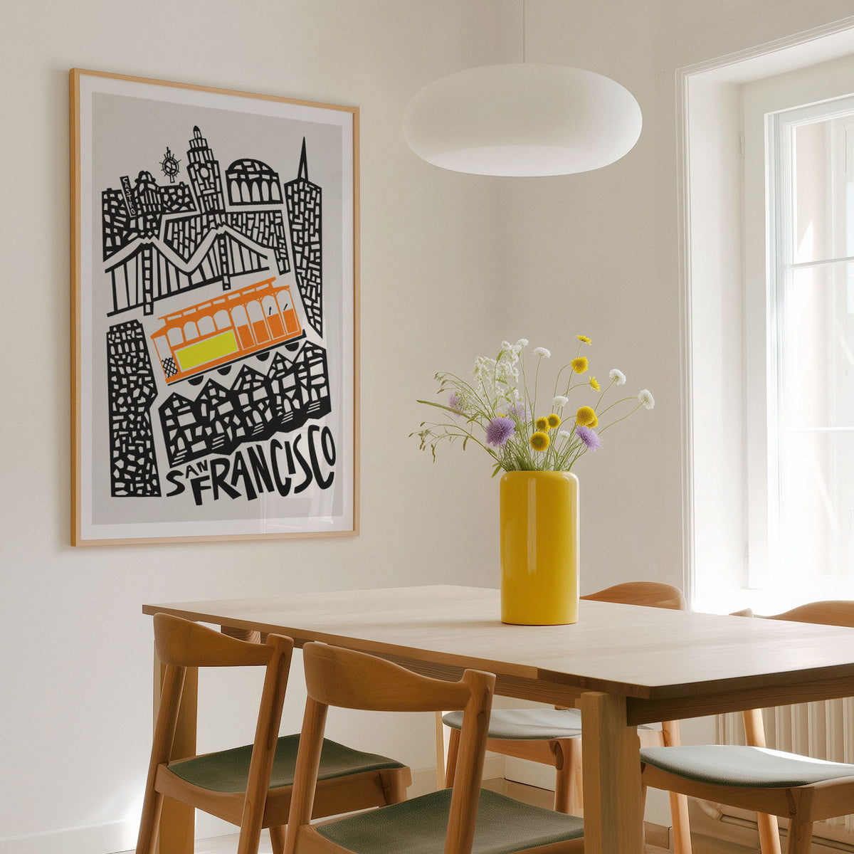 A framed black and white San Francisco cityscape art print featuring an orange cable car, hanging on a light beige wall in a modern dining room with a wooden table, chairs, and a yellow vase with flowers.