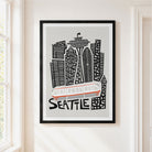 Framed Seattle cityscape wall art hanging on a white wall, featuring black and gray building silhouettes, a white and orange transit vehicle, and the word SEATTLE in black. The frame is black and the print is shown in a bright interior setting.