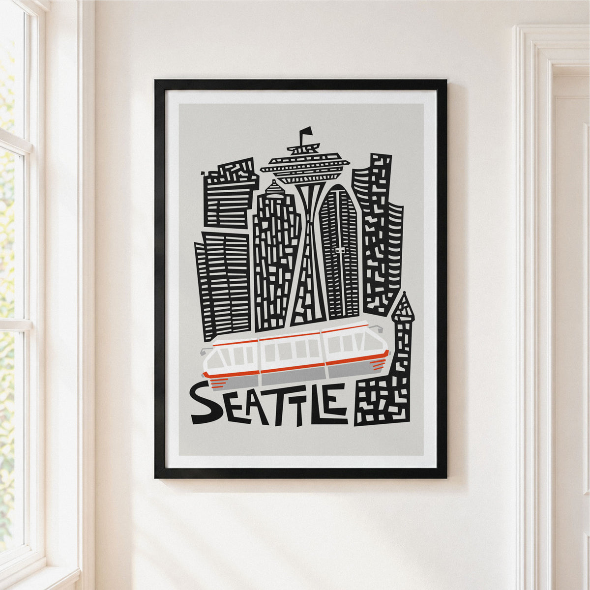 Framed Seattle cityscape wall art hanging on a white wall, featuring black and gray building silhouettes, a white and orange transit vehicle, and the word SEATTLE in black. The frame is black and the print is shown in a bright interior setting.