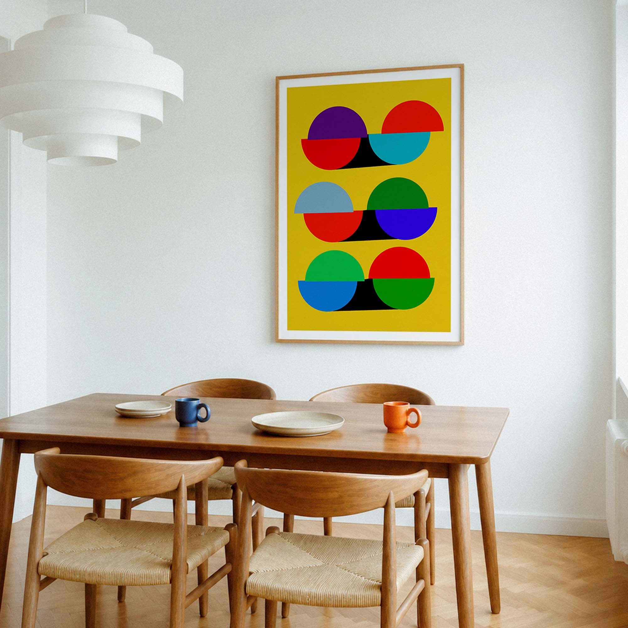 A framed abstract art print with colorful geometric shapes on a yellow background, hanging on a white wall above a wooden dining table and chairs in a bright room.