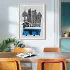 A framed art print featuring a black and blue Seoul cityscape illustration with skyscrapers and a bus, hanging on a light gray wall in a dining room setting with wooden chairs and a table.