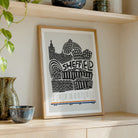 A framed art print featuring a black and white graphic illustration of Sheffield landmarks with the word 'SHEFFIELD' in the center, styled on a light wood shelf next to decorative ceramic vases.
