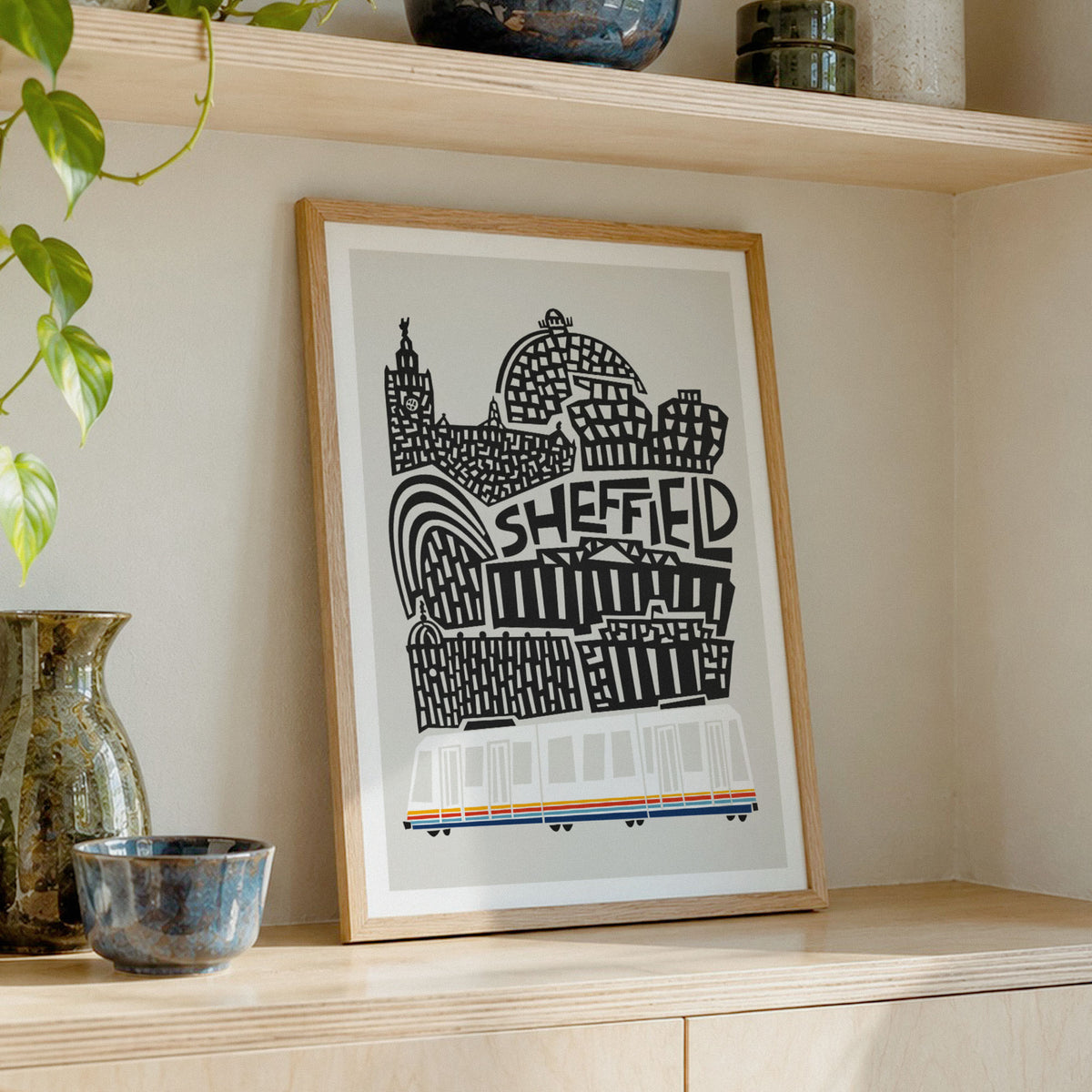 A framed art print featuring a black and white graphic illustration of Sheffield landmarks with the word 'SHEFFIELD' in the center, styled on a light wood shelf next to decorative ceramic vases.