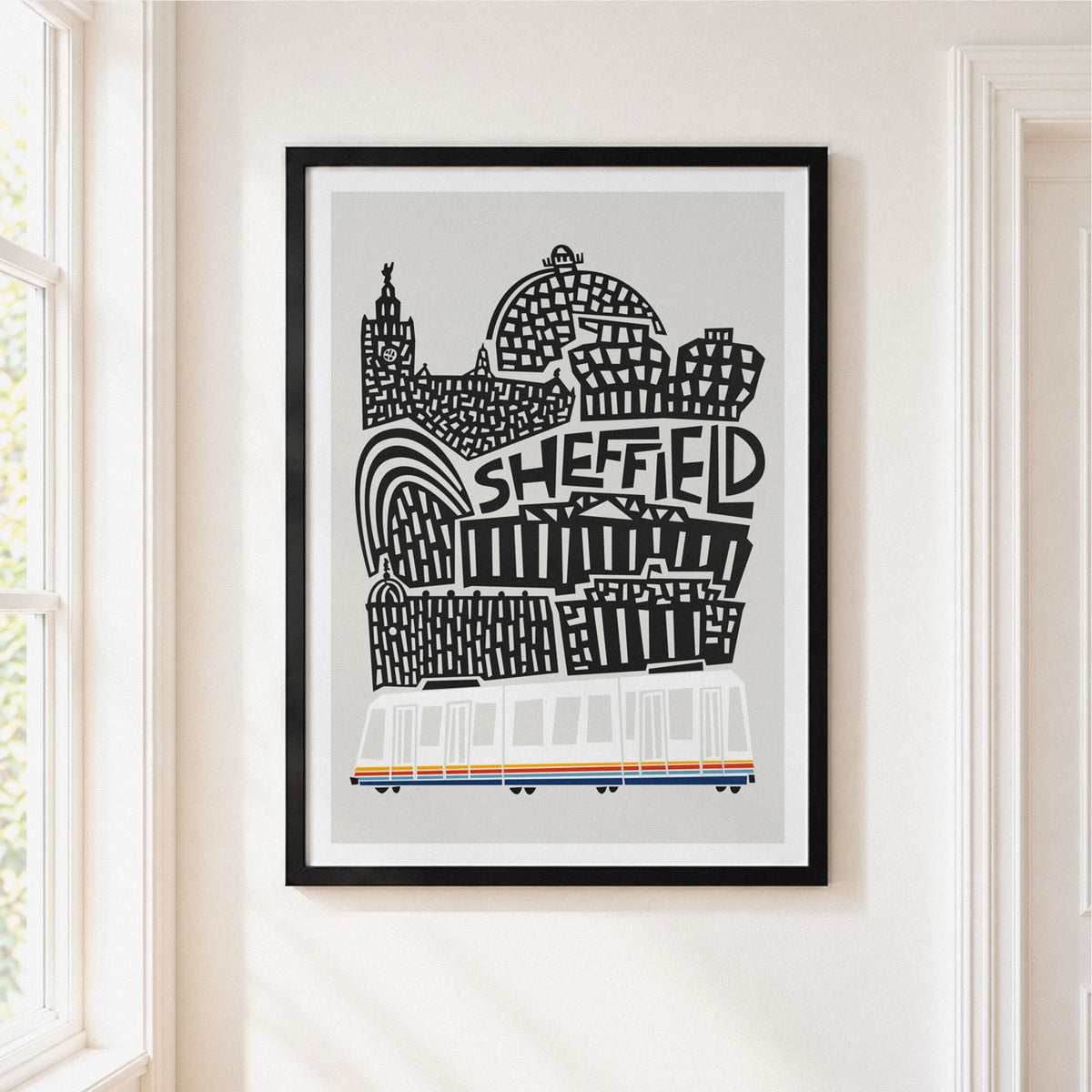 A framed art print featuring a black and white hand-drawn illustration of Sheffield landmarks and a white tram with orange and blue stripes, hanging on a cream-colored wall next to a window.