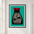 A framed art print featuring a stylized soy sauce bottle with a red cap and cream label on a teal background, hung on a white wall next to a window.