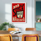A framed art print featuring a retro-style illustration of a margarita glass with the words 'SPICY MARGARITA' on a red background, hanging on a light gray wall in a dining room setting with wooden chairs and a table.