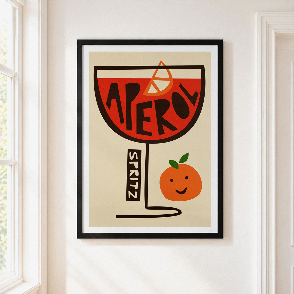 A framed art print featuring a stylized orange cocktail glass with the word  in black over a red-orange liquid, plus a smiling orange character and a vertical 'SPRITZ' label, displayed on a white wall near a window.