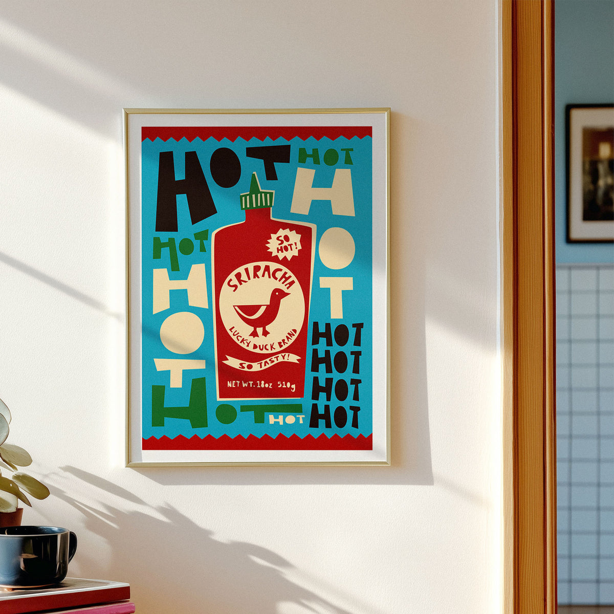A framed art print featuring a retro hot sauce bottle design with the word 'SKIRACH' and repeated 'HOT' text, hanging on a white wall in a real interior setting with sunlight and a plant visible.