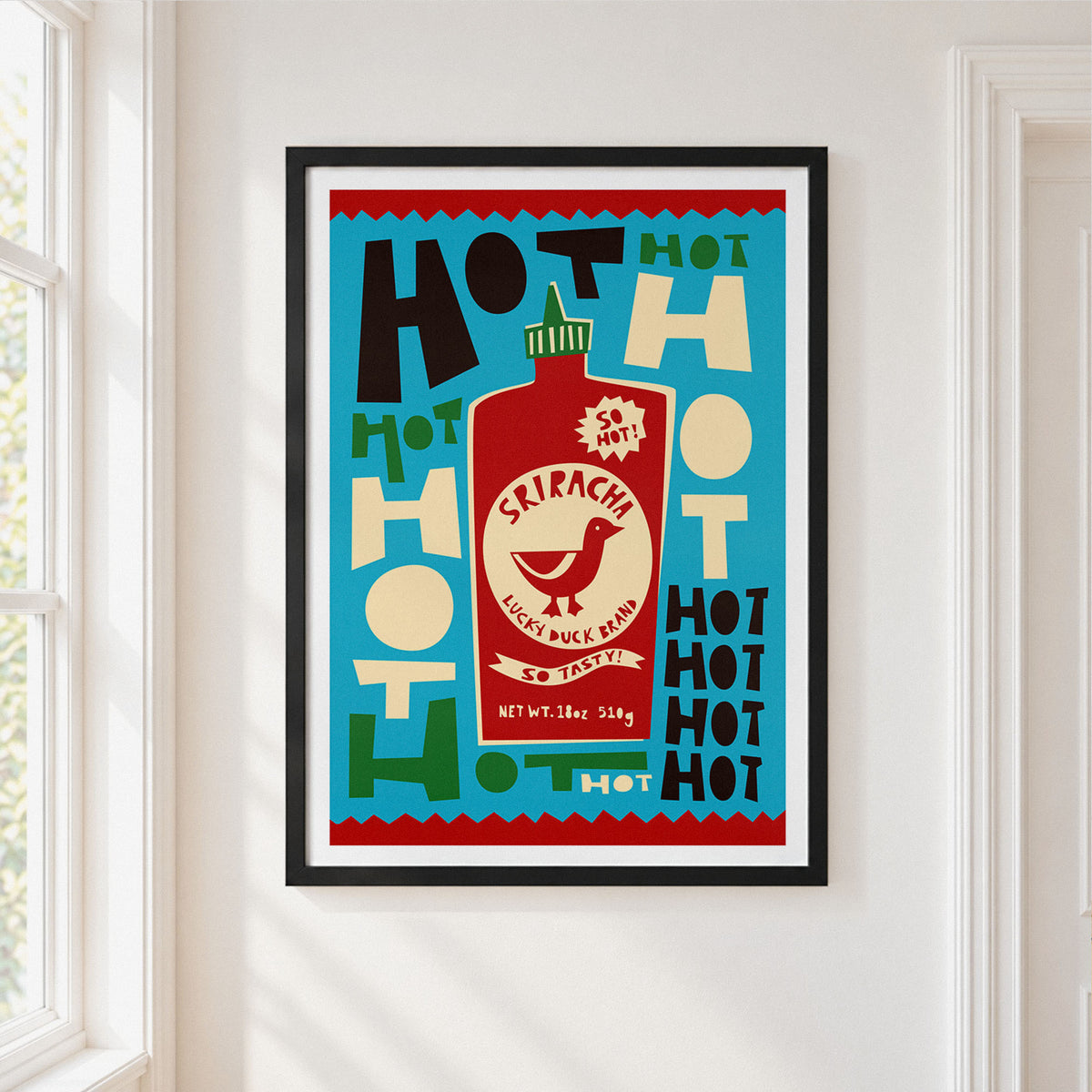 A framed art print featuring a retro Sriracha hot sauce bottle design, hung on a white wall next to a window.