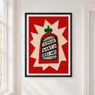 Framed wall art featuring a stylized hot sauce bottle with the words 'SRIRACHA HOT VERY HOT' on a red and cream background, hung on a white wall next to a window.