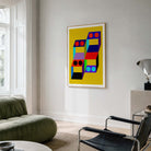 A framed abstract geometric wall art print with a bright yellow background and colorful block shapes hangs on a white wall in a modern living room setting with a green sofa and black chair.