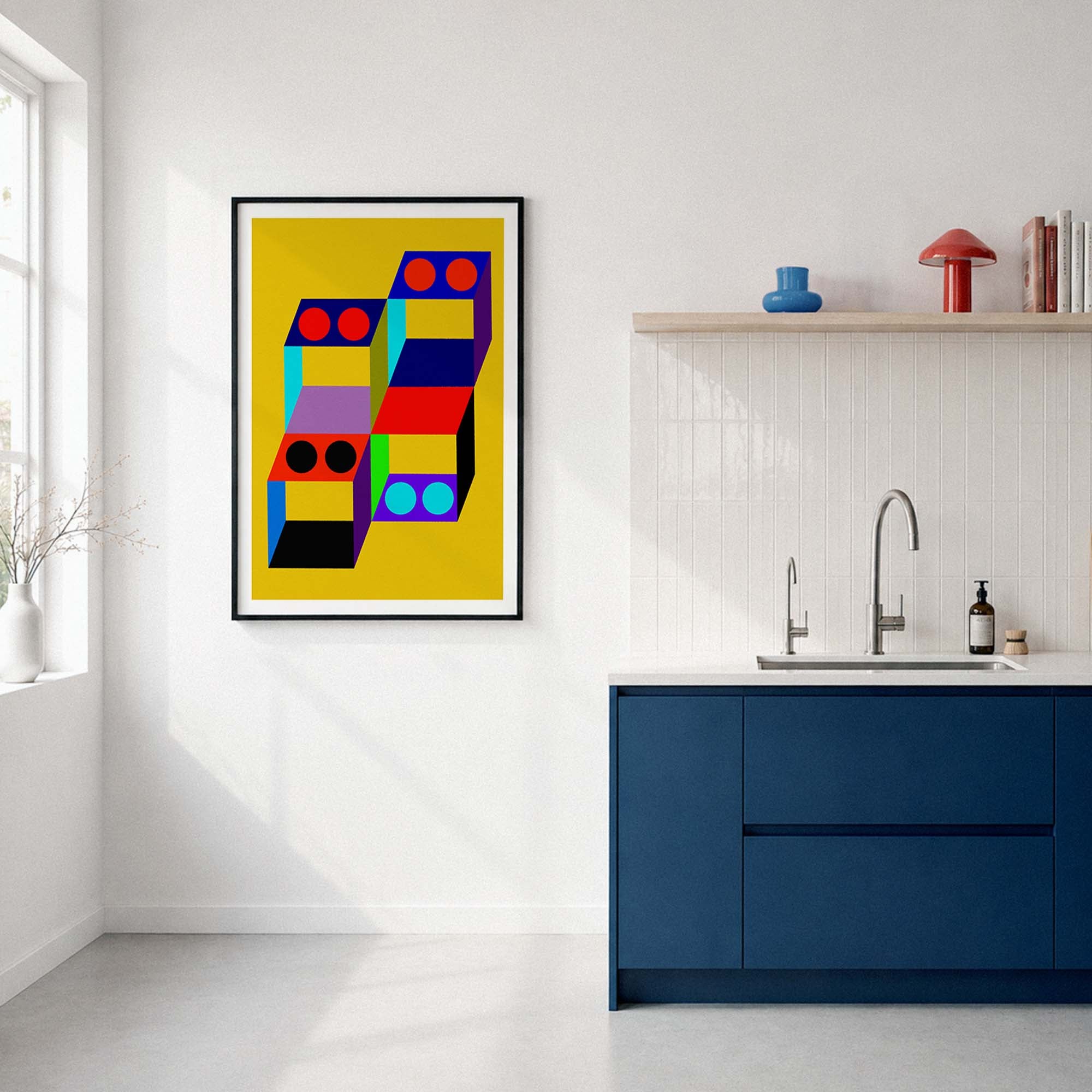 A framed abstract geometric art print with a bright yellow background and colorful 3D block shapes hangs on a white wall in a modern kitchen setting with a blue cabinet and sink.