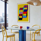 A framed abstract geometric art print with a bright yellow background and colorful stacked block shapes hangs on a white wall in a modern dining room with a glass table, yellow chairs, and a wicker pendant light.