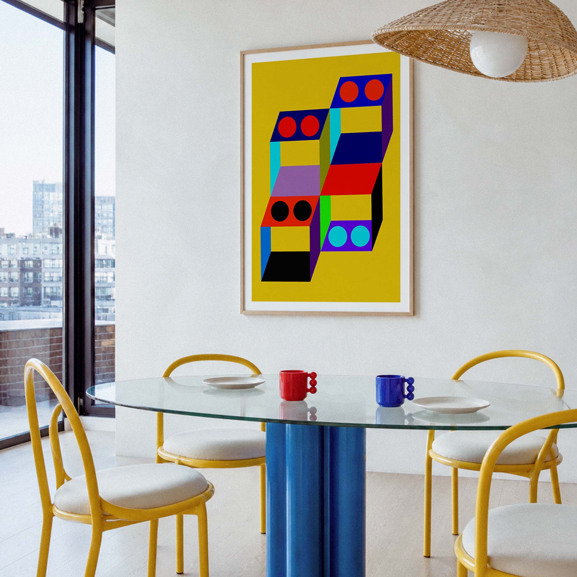 A framed abstract geometric art print with a bright yellow background and colorful stacked block shapes hangs on a white wall in a modern dining room with a glass table, yellow chairs, and a wicker pendant light.