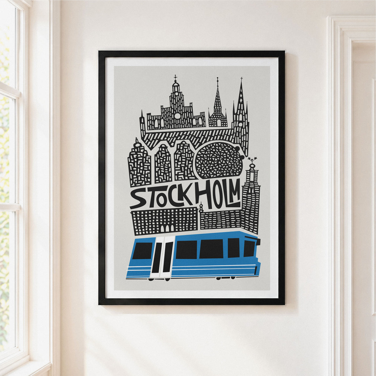 A framed art print featuring a stylized Stockholm cityscape with a blue tram and the word 'STOCKHOLM' in black, hung on a light beige wall next to a window.