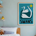 A framed wall art print featuring two white swans and the word “SWANS” in black, hanging on a light blue-gray wall above a bed in a styled bedroom.