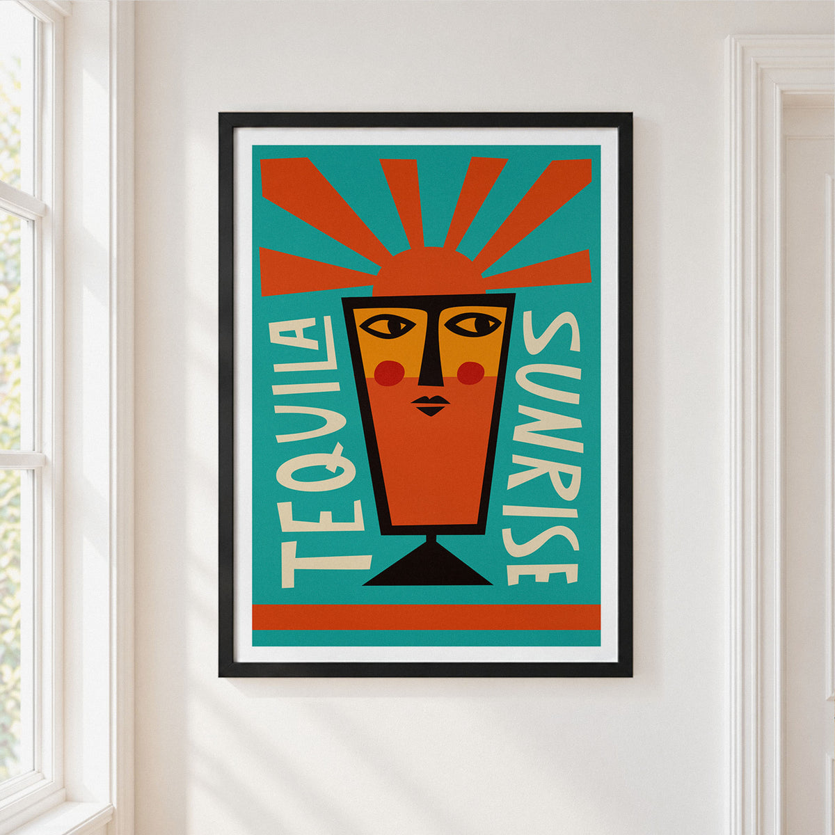A framed art print featuring a stylized orange tequila glass with a face and radiating orange sun rays on a teal background, with the words 'TEQUILA SUNRISE' in white. The print is displayed on a white wall next to a window.