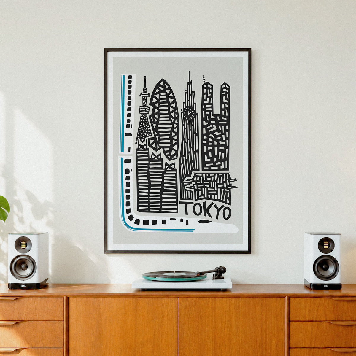 A framed black and white Tokyo cityscape art print with light blue accents hangs on a white wall above a wooden credenza, flanked by two white speakers and a vinyl record player.
