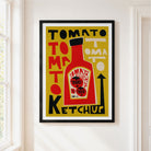 A framed pop-art style poster featuring a red ketchup bottle and bold text reading 'TOMATO KETCHUP' on a mustard yellow background, hung on a white wall next to a window.