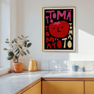A framed art print featuring a stylized red tomato and the word 'TOMATO' in bold pink and cream lettering on a black background, hanging on a white kitchen wall above a wooden counter with a potted plant and mugs nearby.