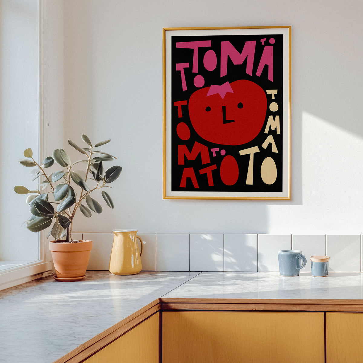 A framed art print featuring a stylized red tomato and the word 'TOMATO' in bold pink and cream lettering on a black background, hanging on a white kitchen wall above a wooden counter with a potted plant and mugs nearby.