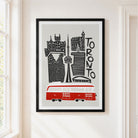 A framed art print featuring a stylized black and white illustration of the Toronto skyline with a red transit bus, hanging on a cream-colored wall next to a window.