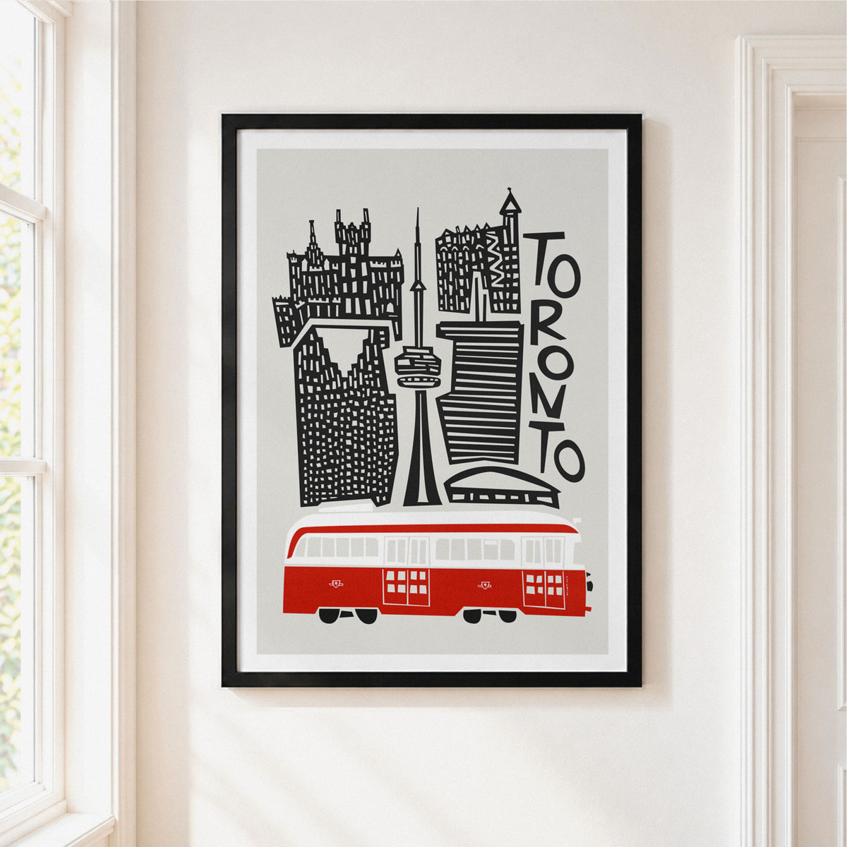 A framed art print featuring a stylized black and white illustration of the Toronto skyline with a red transit bus, hanging on a cream-colored wall next to a window.