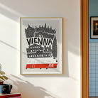 A framed art print featuring a black and white illustration of Vienna landmarks with a red tram, hanging on a light gray wall in a modern dining room setting.