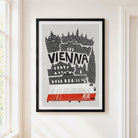 A framed art print featuring a stylized black and white illustration of Vienna with a red tram, hanging on a light beige wall next to a window.