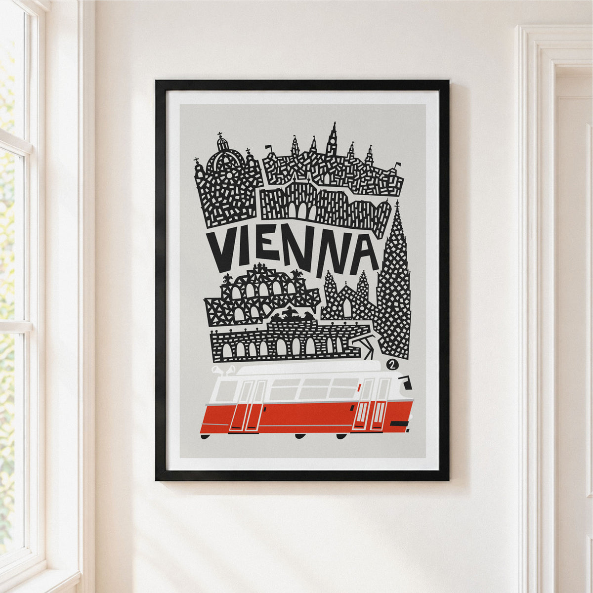 A framed art print featuring a stylized black and white illustration of Vienna with a red tram, hanging on a light beige wall next to a window.