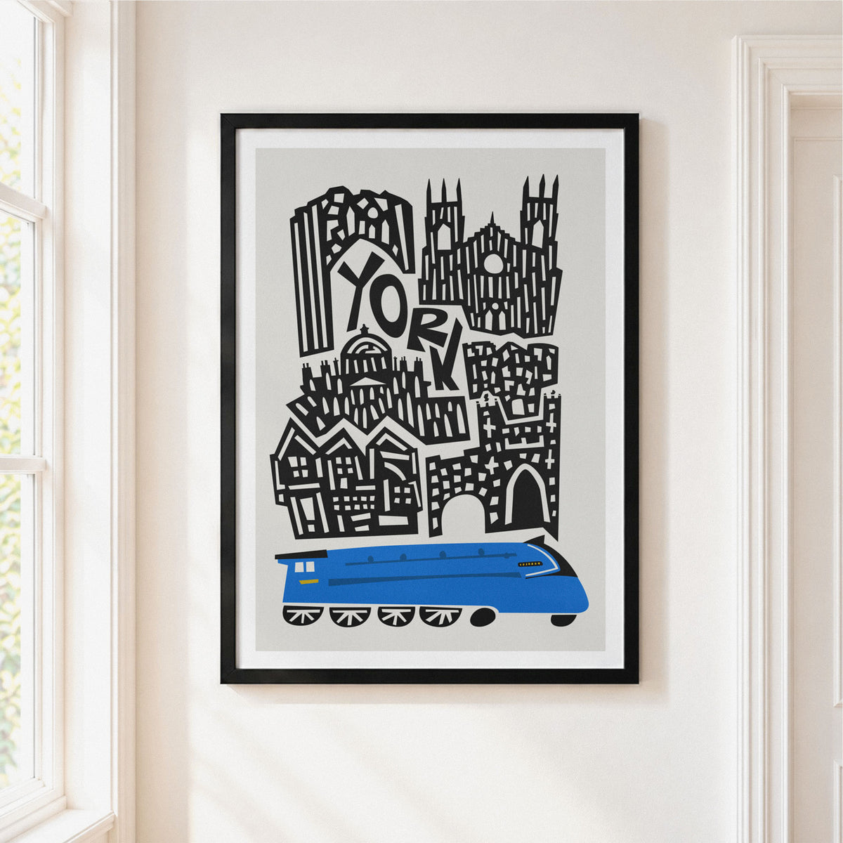 A framed art print featuring a stylized black and white illustration of York with a bright blue train at the bottom, hanging on a cream-colored wall next to a window.