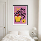 A framed art print featuring a retro-style 'Amaretto Sour' cocktail illustration in pink, yellow, black, and red, displayed on a bedroom wall above a bed.
